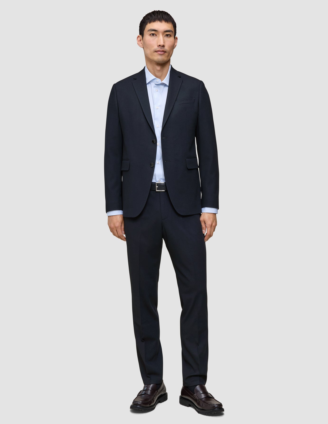 En mann kledd i Motion Suit-bukser i størrelse Regular i fargen Midnight Blue, med en lyseblå skjorte og mørke skinnloafers.
